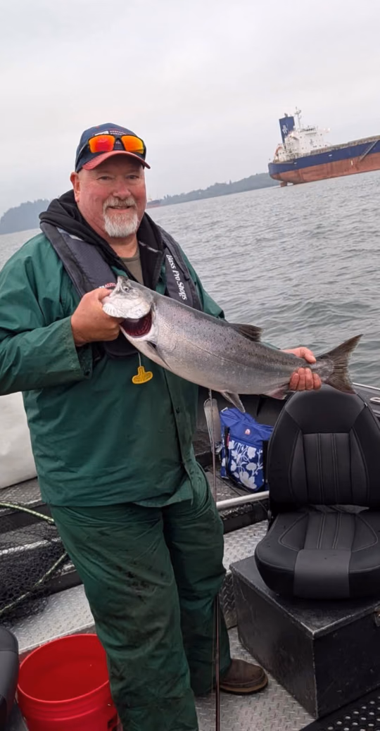 Angler in green rain gear holding a large fish, with a cargo ship on the water behind