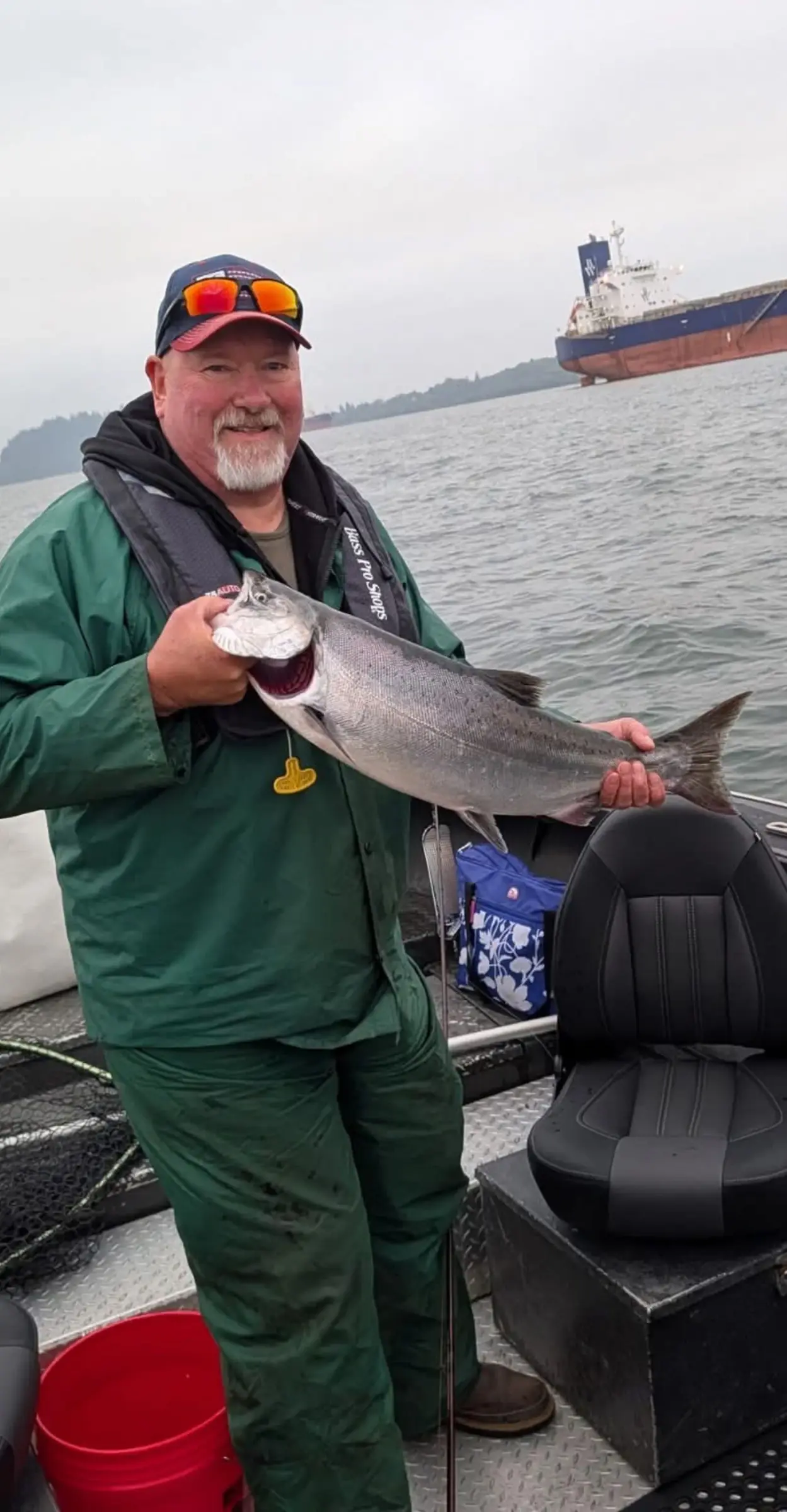 Angler in green rain gear holding a large fish, with a cargo ship on the water behind