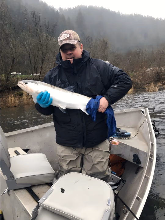 Angler in a camouflage cap holding a bright fish with a blue glove, boat and riverbank behind