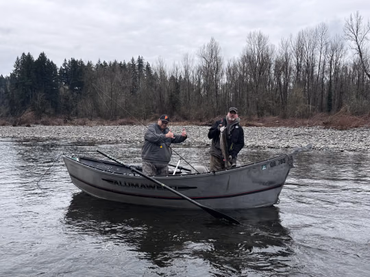 Two anglers standing in an Alumaweld drift boat on a wide river, one holding a fish