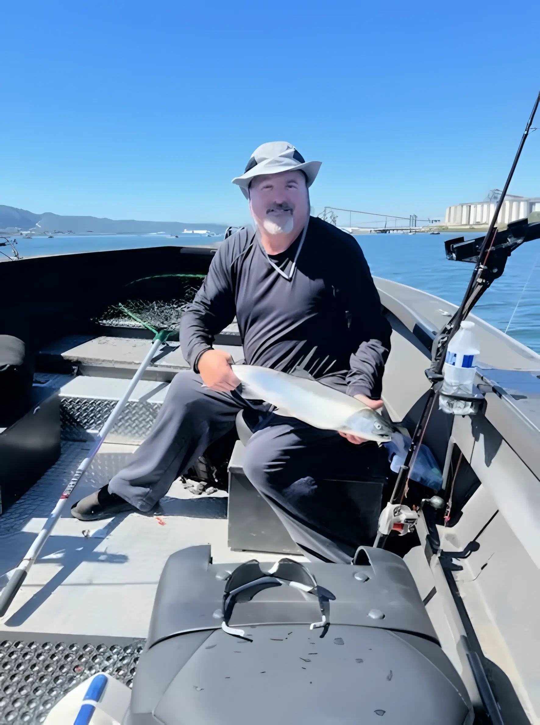 Angler in a bucket hat seated on a boat holding a fish, a bridge in the distance