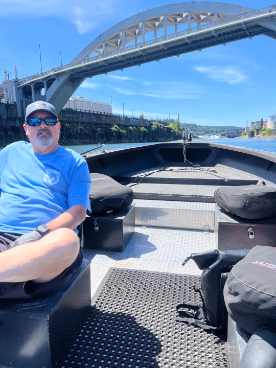 Angler seated on the open deck of a sled boat passing under a steel arch bridge