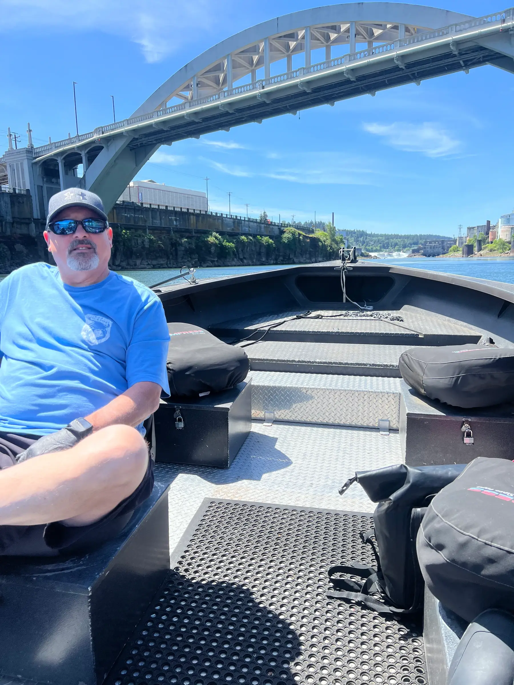 Angler seated on the open deck of a sled boat passing under a steel arch bridge