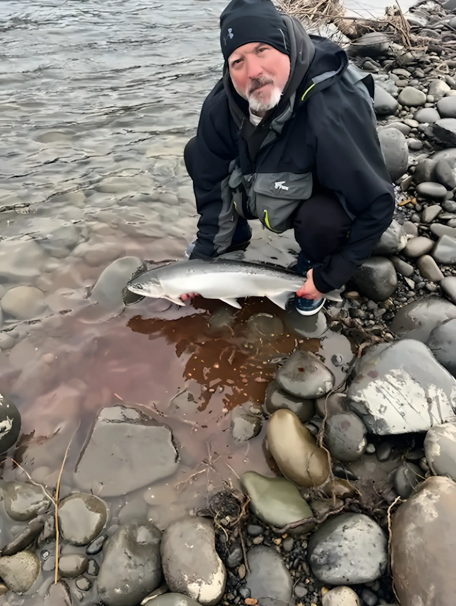 Angler in a dark jacket crouching on a rocky riverbank with a silver fish