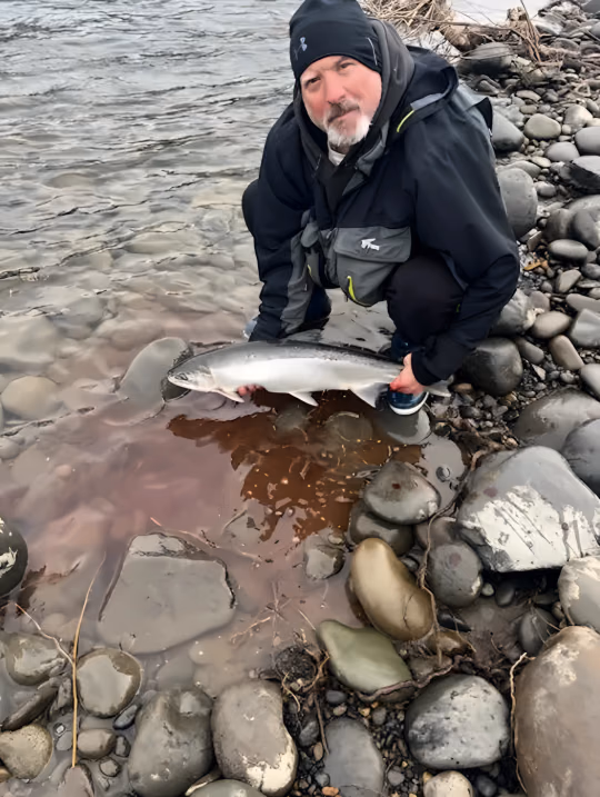 Angler in a dark jacket crouching on a rocky riverbank with a silver fish