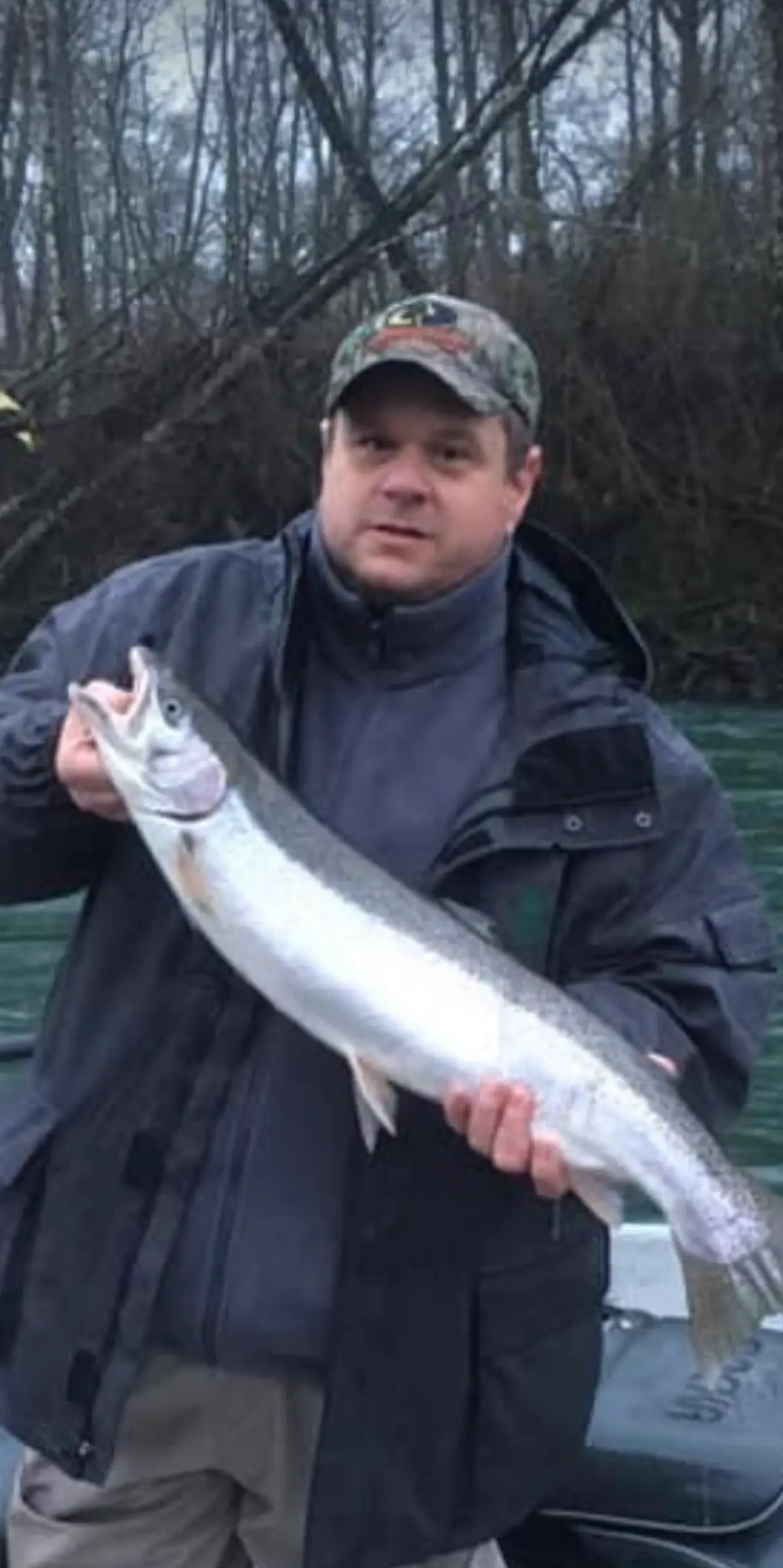 Angler in a camo cap holding a fish, bare winter trees behind
