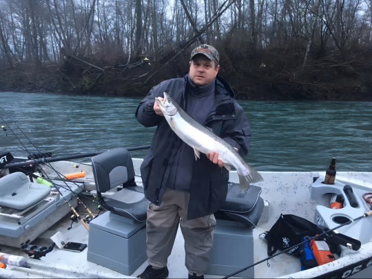 Angler on a boat holding a fish, bare trees on the riverbank behind