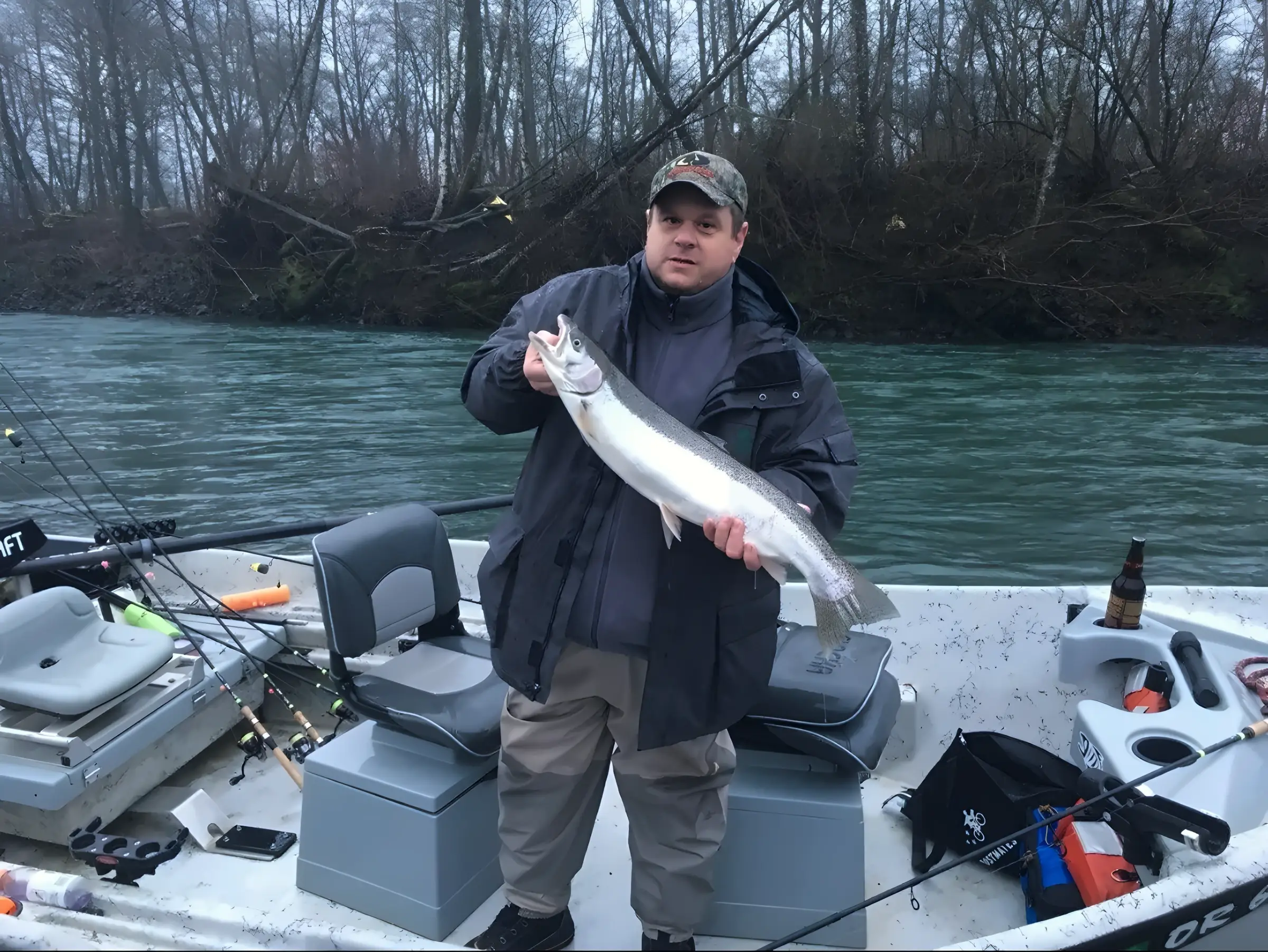 Angler on a boat holding a fish, bare trees on the riverbank behind